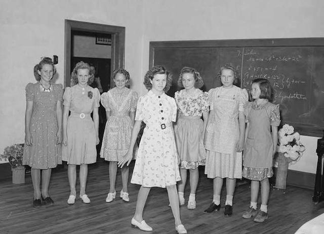 A group of young girls proudly showcasing their handmade dresses in a Home Economics class, demonstrating the sewing skills they acquired.